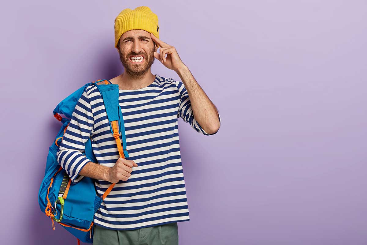 Stressful male adventurer has headache after tiring trip, clenches teeth from pain, wears stylish outfit, poses with backpack, cannot concentrate, poses over purple wall, free space for your advert