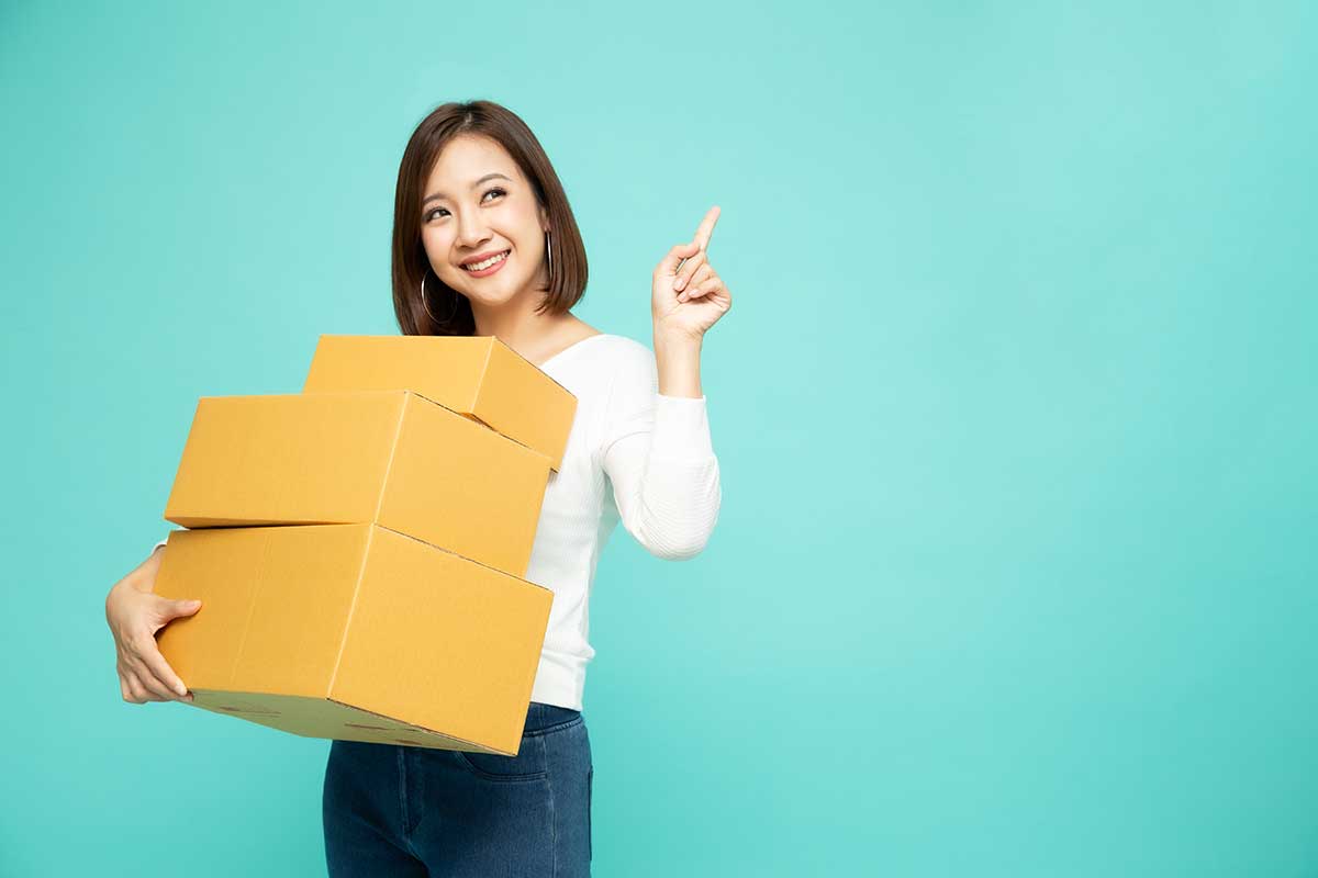 Happy Asian woman holding package parcel box and pointing finger to empty copy space isolated on light green background, Delivery courier and shipping service concept