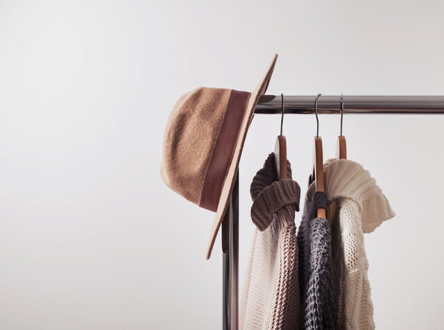 Knitted sweaters on hangers and felt hat on the white wall background. Autumn or winter wardrobe.